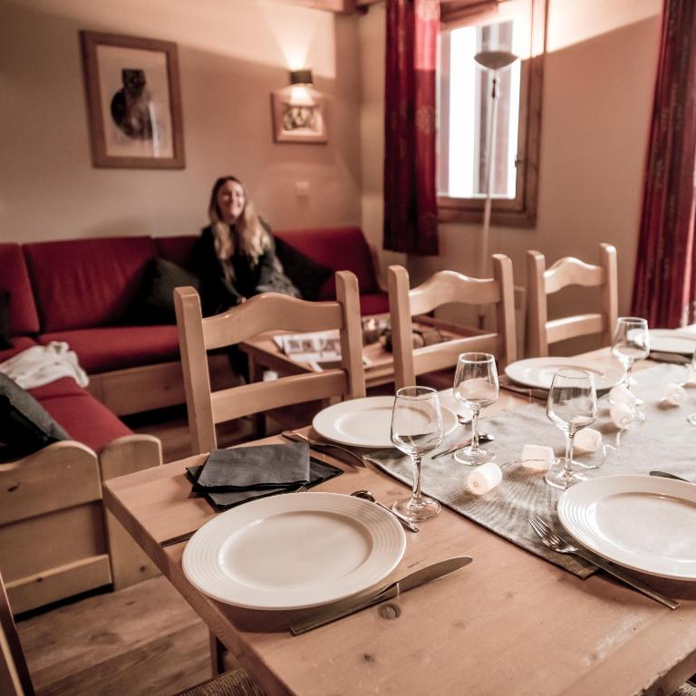 Mūsdienīga viesistaba ar ēdamgaldu, kas klāts ar baltām šķīvjiem un glāzēm, aizmugurē sarkana dīvāna un sievietes siluets. Ideāla vide mājīgām ģimenes vakariņām.