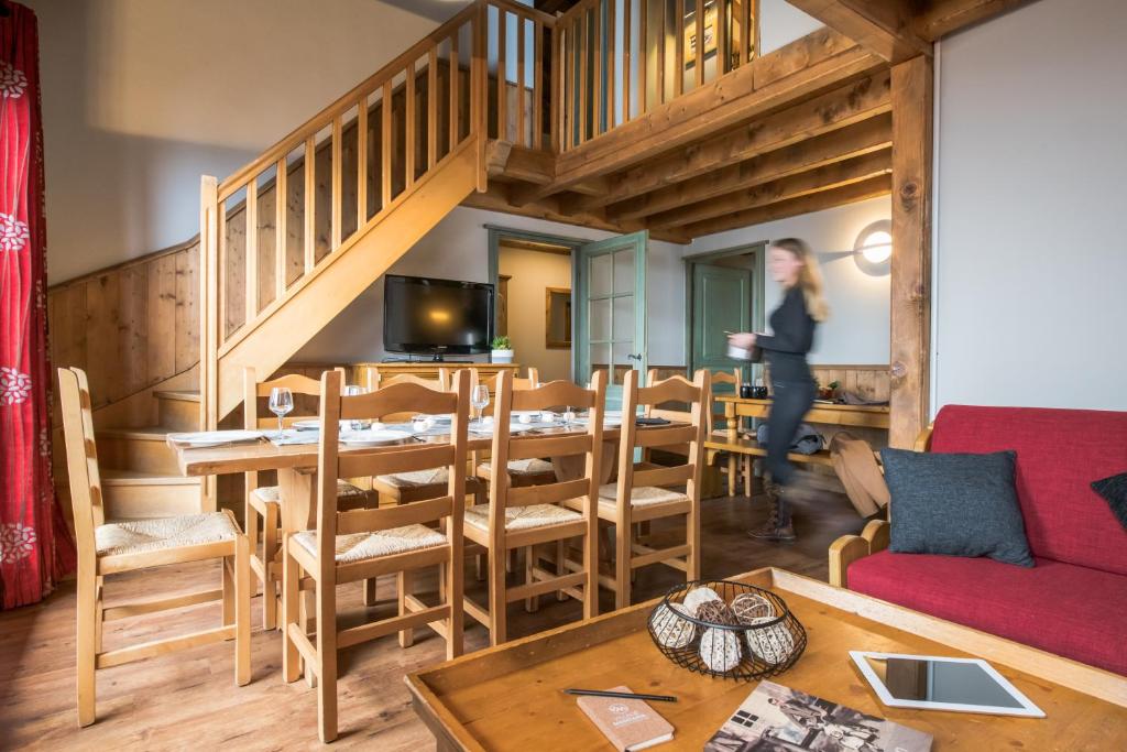 Mūsdienīga viesistaba ar koka mēbelēm, ēdamgaldu un kāpnēm uz otro stāvu, kurā redzama sieviete, kas nes kafiju. Interjerā ir siltas krāsas un mājīga atmosfēra.