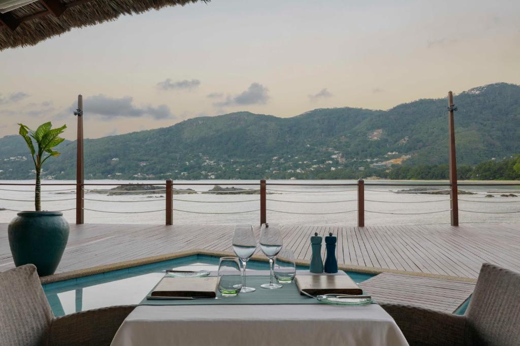 Skats uz mierīgu ūdeni un kalniem no eleganta restorāna terases, kur uz galda ir glāzes un galda piederumi. Ideāla vieta romantiskām pusdienām vai vakariņām.