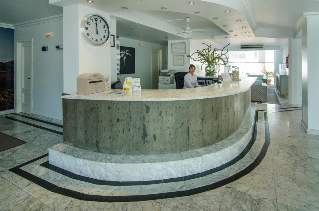 Recepcija ar modernu dizainu, kurā strādā darbinieks, un redzama pulksteņa un augu dekorācija. Telpa ir gaiša un plaša, ar marmora grīdu.