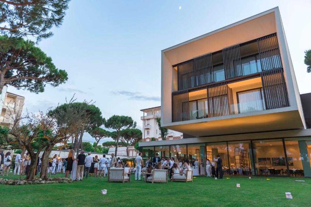 Mūsdienīga māja ar lieliem logiem un terasi, ap kuru norisinās pasākums dārzā, kur cilvēki ģērbušies baltā.