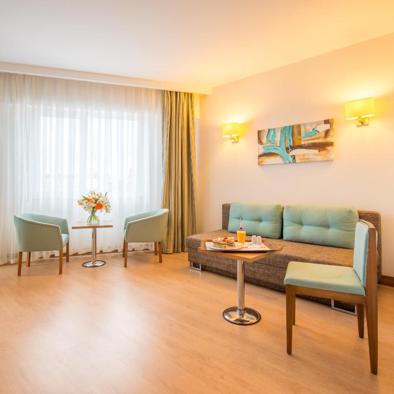 Mūsdienīgs viesnīcas numurs ar gaišu interjeru, kurā ir dīvāns un divas krēsli, kā arī galdiņš ar dzērieniem un ziediem. Logi ar aizkariem nodrošina dabīgu apgaismojumu.