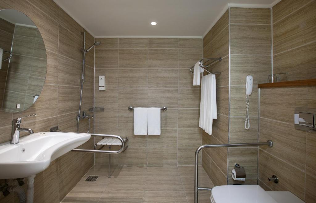 Mūsdienīga vannas istaba ar gaismu brūnu flīžu apdari, aprīkota ar dušu, izlietni un piekļūšanas atbalsta sistēmām. Vannas istabā ir arī spogulis un dvieļu turētājs.