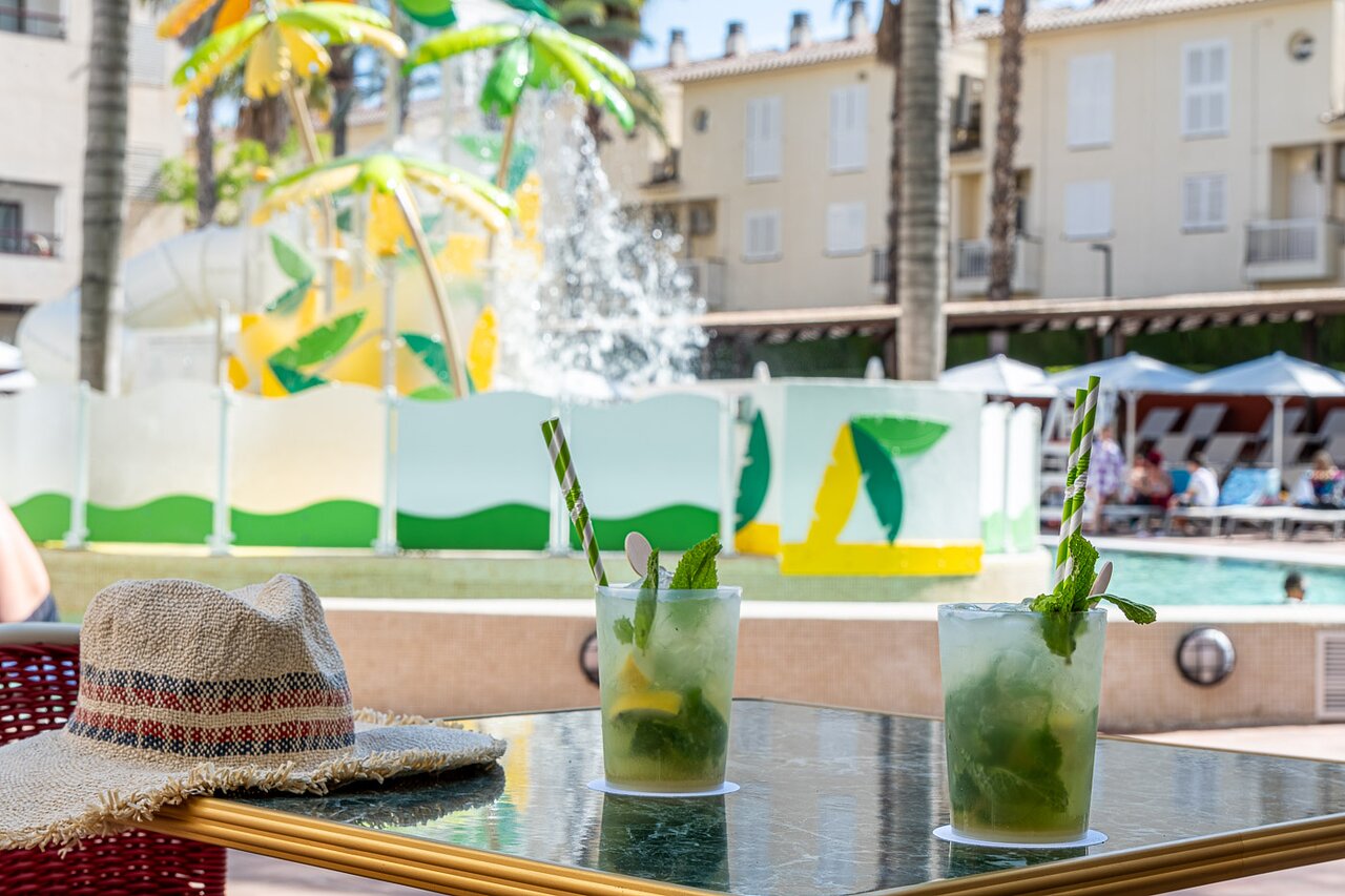Atsvaidzinoši kokteiļi un salmu cepure uz galda pie baseina viesnīcā Estival Park Marena.