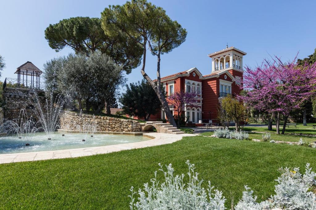 Skaista vēsturiska villa ar sarkanu fasādi, ieskauta zaļā dārzā ar ziediem un strūklaku. Dārzā redzami lieli koki un atpūtas zona.