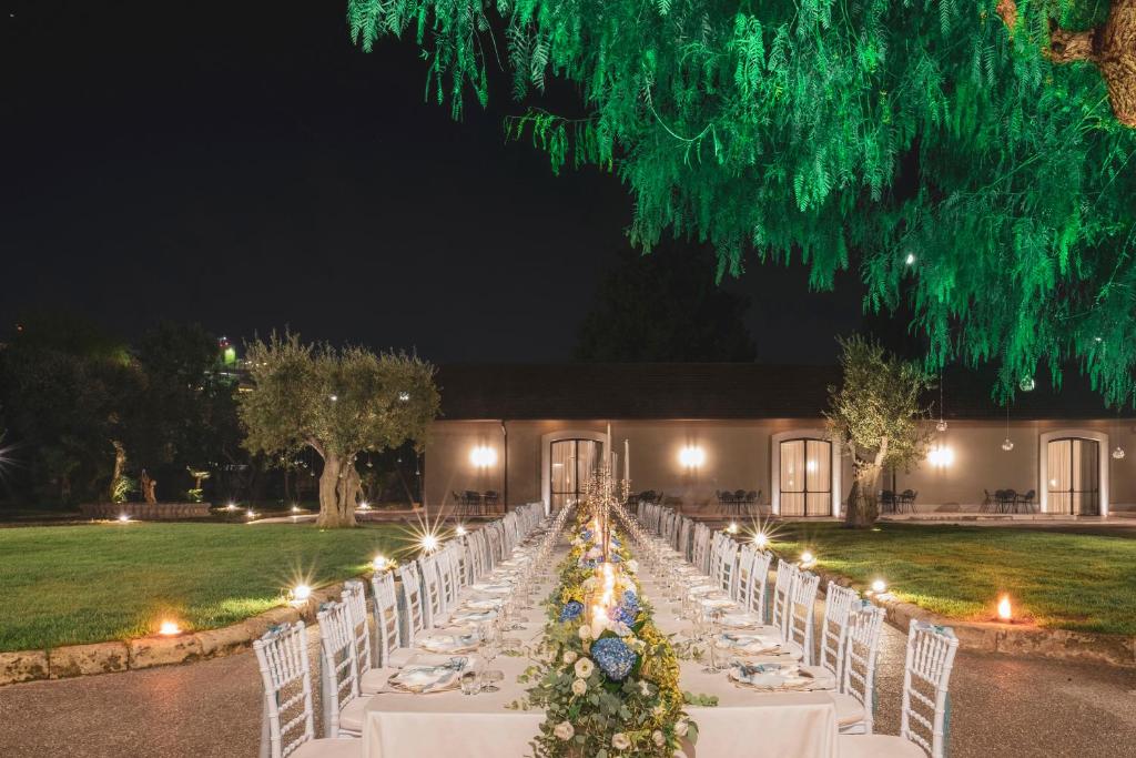 Elegantais vakara galda iestatījums dārzā, apgaismots ar mīkstu gaismu un dekorēts ar ziediem, radot romantisku atmosfēru. Fons veido ainava ar kokiem un ēku, kas piešķir burvīgu noskaņu.