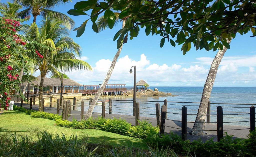 Tropu ainava ar palmu kokiem un ziediem, kas apņem pludmales promenādi pie jūras. Skats uz koka piestātni un mierīgu ūdeni zem zila debesīm.