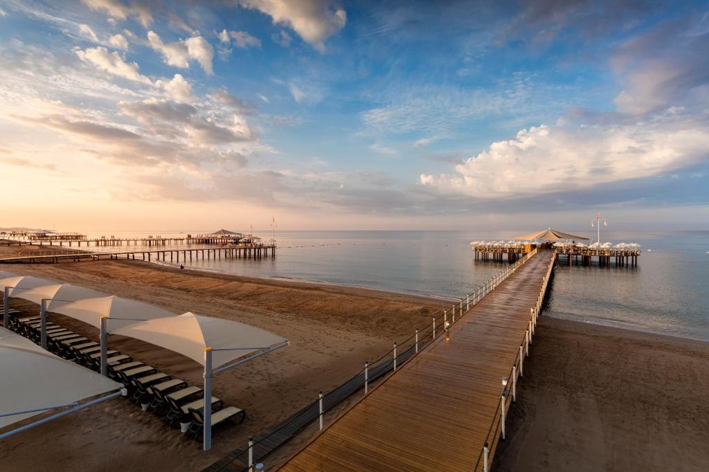 Klusais pludmales skats ar koka piestātni, kas stiepjas uz mierīgu jūru, un mākoņainu debesu fonu. Pludmalē redzami sauļošanās krēsli zem baltiem nojumes.