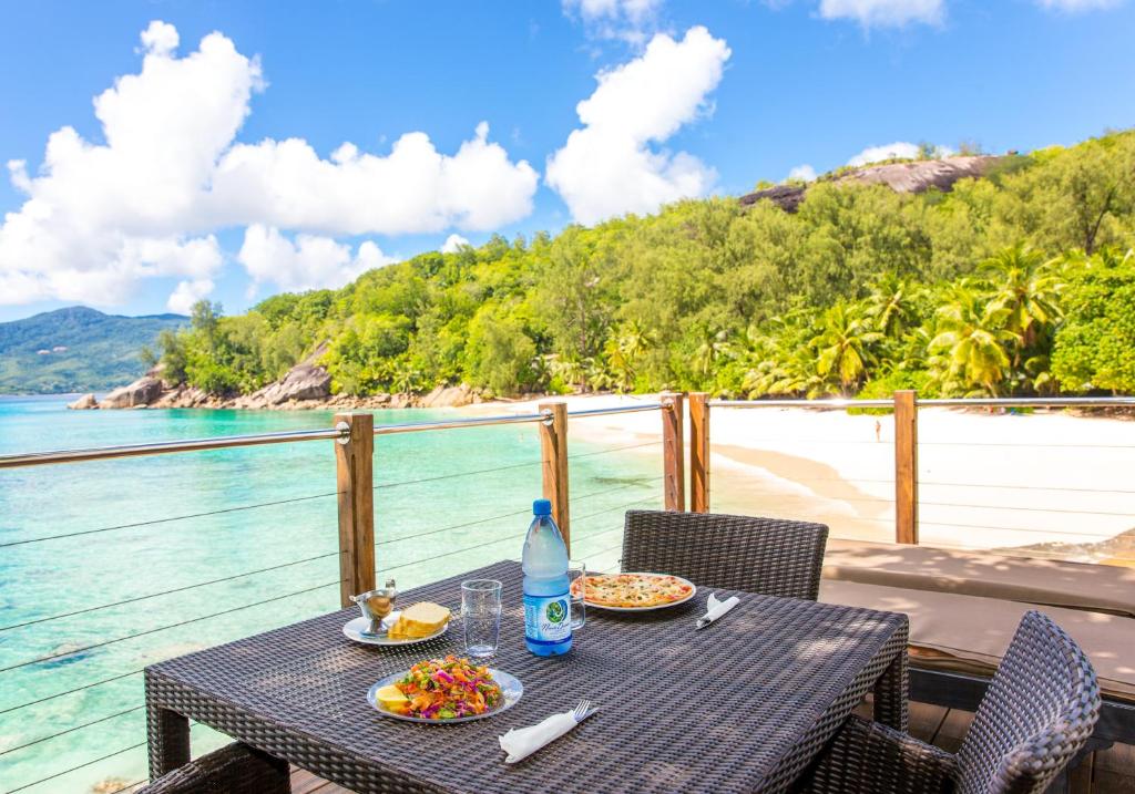 Skaista pludmales ainava ar galdu un ēdieniem, kas pasniegti uz terases pie ūdens. Rāmās debesīs redzami balti mākoņi, bet apkārt ir zaļa daba un palmām apaugušas nogāzes.