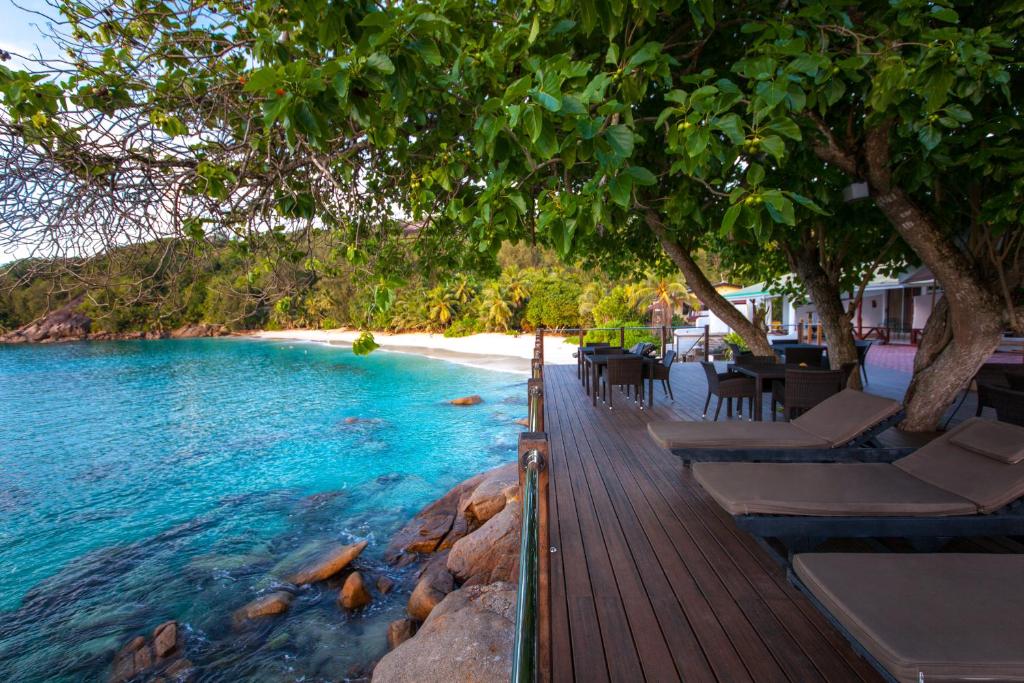 Pludmales kafejnīca ar terasi, kurā ir šūpuļtīkli un galdi, blakus skaidram zilam ūdenim un smilšainai pludmalei. Apkārtējā ainava ir zaļa un tropiska, radot mierīgu atmosfēru.