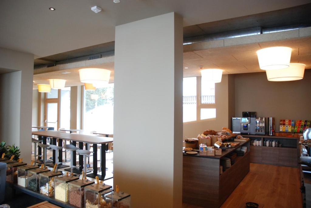 Mūsdienīgs kafijas veikals ar plašu galdu, augstām krēsliem un bagātīgu brokastu izvēli, kas ietver dažādas uzkodas un dzērienus. Gaisma ir silta un aicinoša, radot patīkamu atmosfēru.