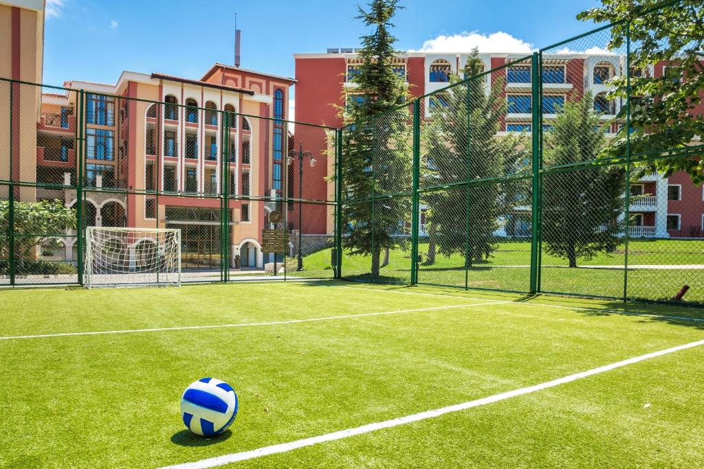 Futbola laukums ar zāles segumu un zilu bumbu, fona redzama moderna ēka un zaļumi.