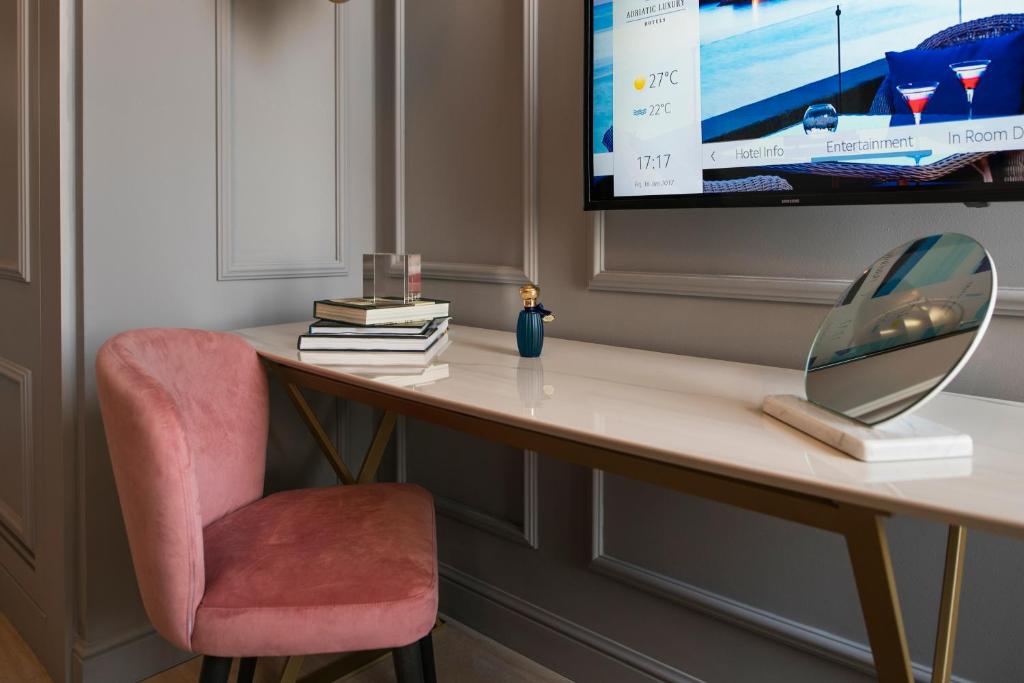 Mūsdienīgs rakstāmgalds ar rozā krāsas krēslu, dekorēts ar grāmatām un spoguli, blakus televizors ar viesnīcas informāciju.