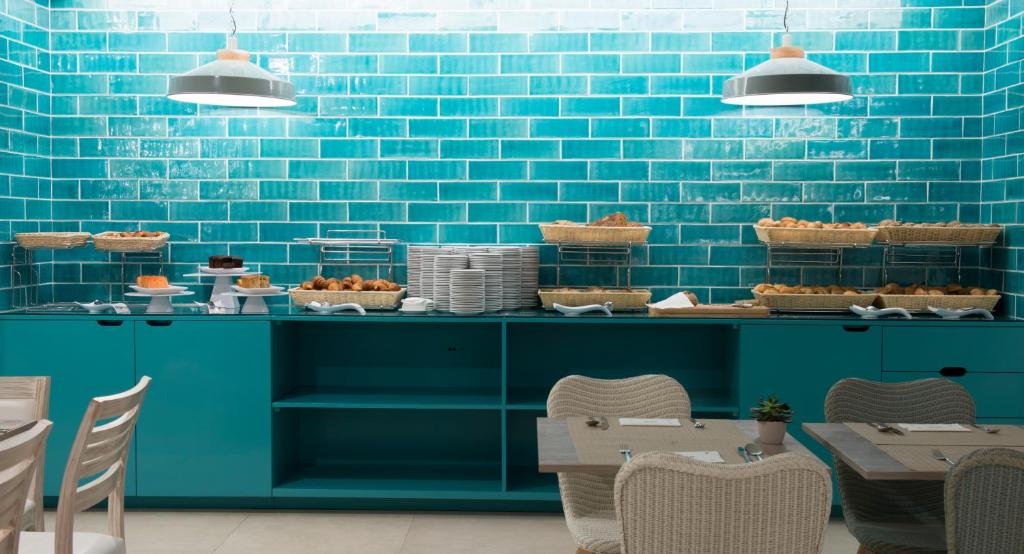 Mūsdienīgs restorāna interjers ar tīrkultūras zilu flīžu sienu, kas izceļ ēdienu galdu ar maizēm un saldumiem. Galdi ir sakārtoti ar elegantiem galda piederumiem un augu dekorāciju.