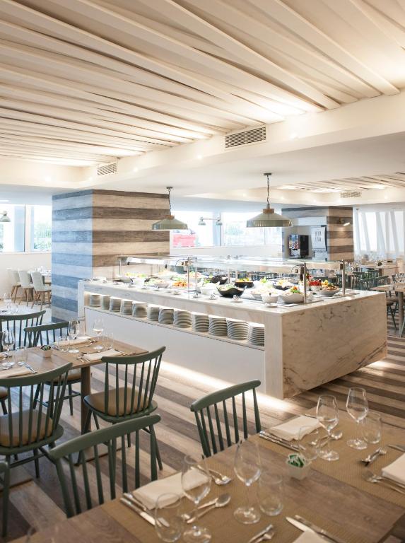 Moderns restorāna interjers ar elegantiem koka galdiem un krēslām. Centrā redzama gaisīga bufete ar dažādiem ēdieniem.