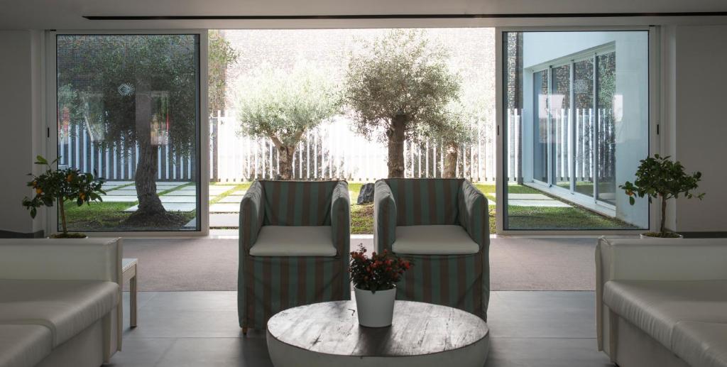 Mūsdienīga telpa ar divām krēslām un galdu, kas vērsta uz dārzu ar olīvkoku un zāles laukumu. Dabas gaisma ienāk caur lielajiem logiem, radot mierīgu atmosfēru.