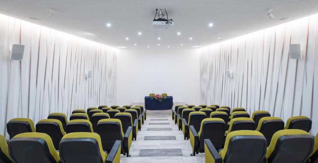 Mūsdienīga konferenču zāle ar dzeltenām un melnām krēslām, izceļas ar elegantu dizainu un labu apgaismojumu. Zāles priekšpusē ir skatuves zona ar galdu un ziediem.