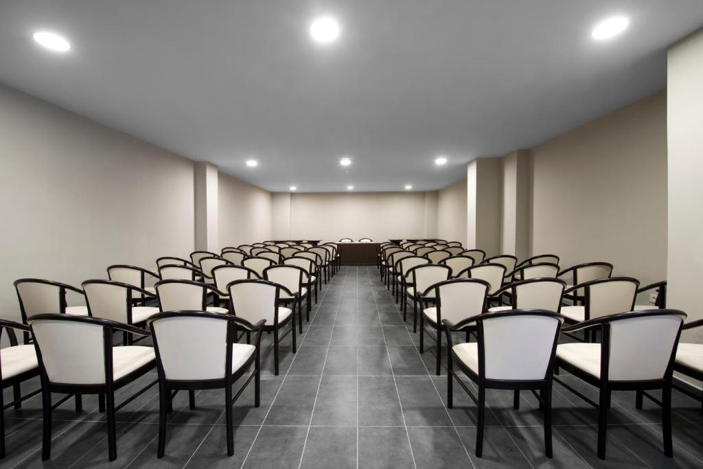 Konferenču zāle ar rindām krēsliem un neitrālu interjeru, piemērota sanāksmēm un pasākumiem. Apgaismojums rada gaišu un plašu atmosfēru.