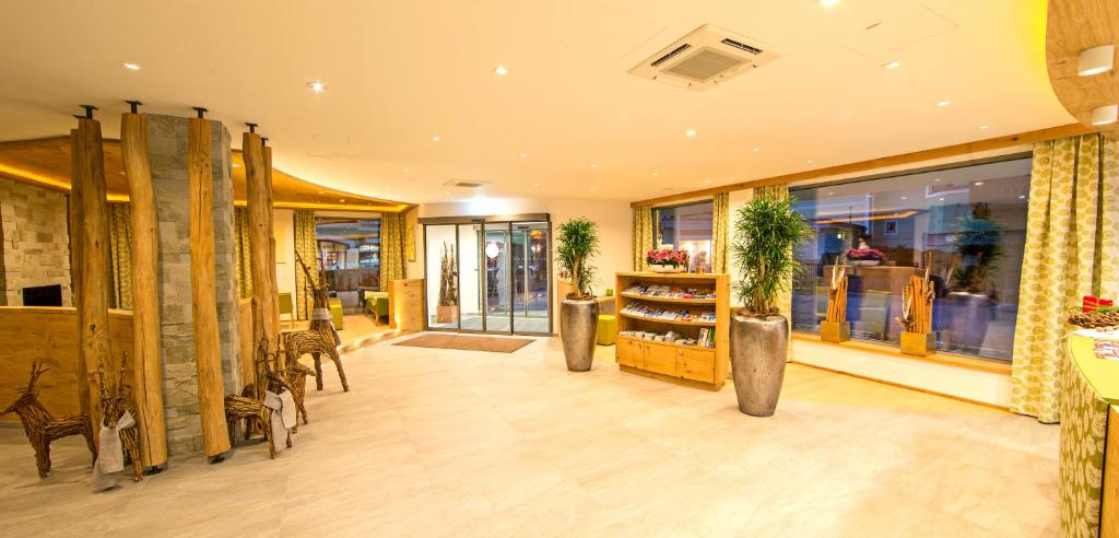 Mūsdienīgs viesnīcas interjers ar koka un akmens elementiem, plaša ieeja un dabiska apgaismojuma izmantošana. Interjerā ir redzami augi un dekoratīvi priekšmeti, kas rada siltu un viesmīlīgu atmosfēru.