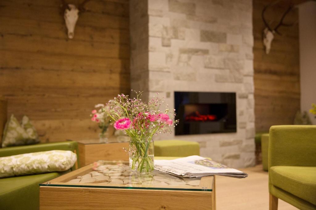 Mūsdienīgs interjers ar zaļām krēsliem un koka elementiem, uz galda novietota ziedu pušķis un avīze, fons ar akmens sienu un mākslīgo kamīnu.