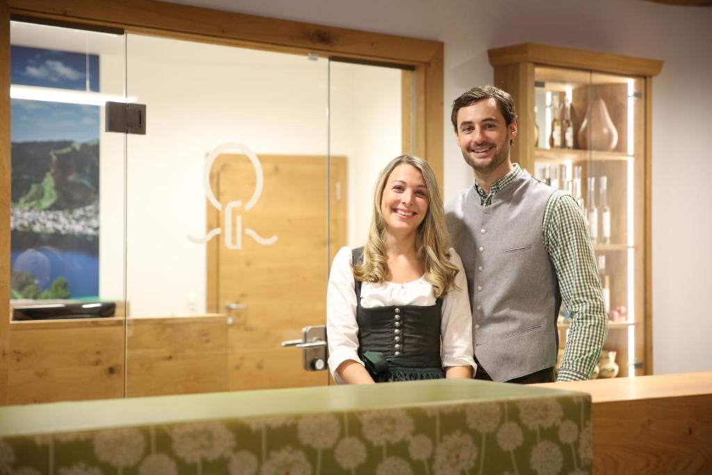 Smaidoši viesmīļi stāv pie reģistratūras, ģērbušies tradicionālās tērpā, ar koka fona un modernām interjera detaļām.