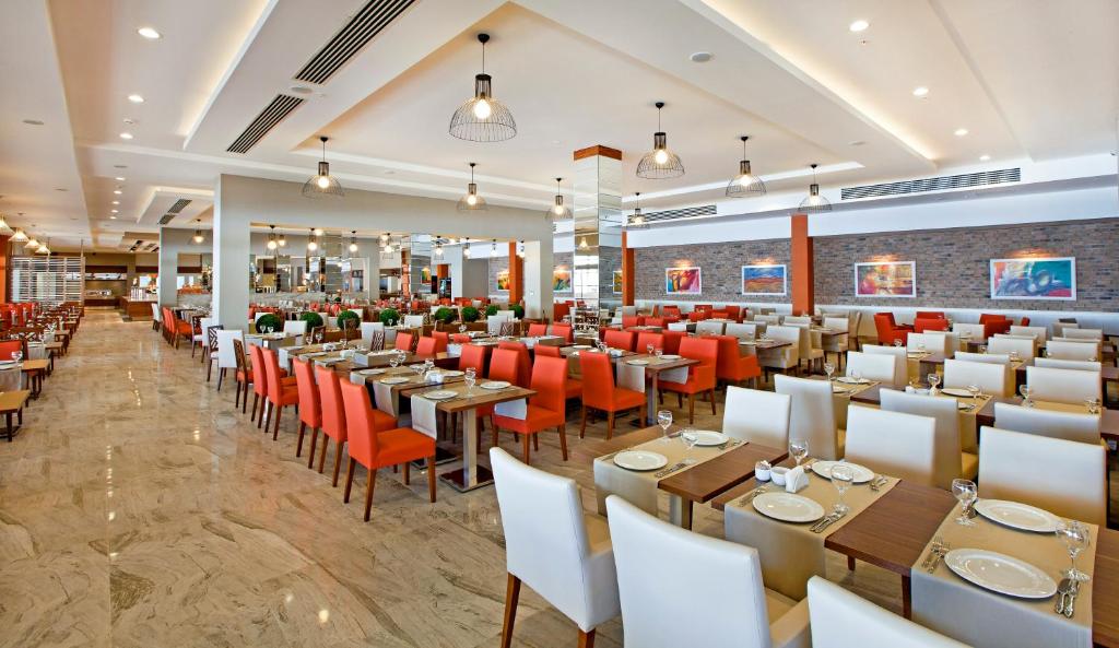 Mūsdienīga restorāna iekšskats ar elegantiem galdiem un krēsliem, kas izkārtoti plašā telpā. Sarkanā un baltā krāsā veidotie krēsli piešķir atmosfērai dzīvīgumu, bet gaismas elementi rada patīkamu noskaņu.