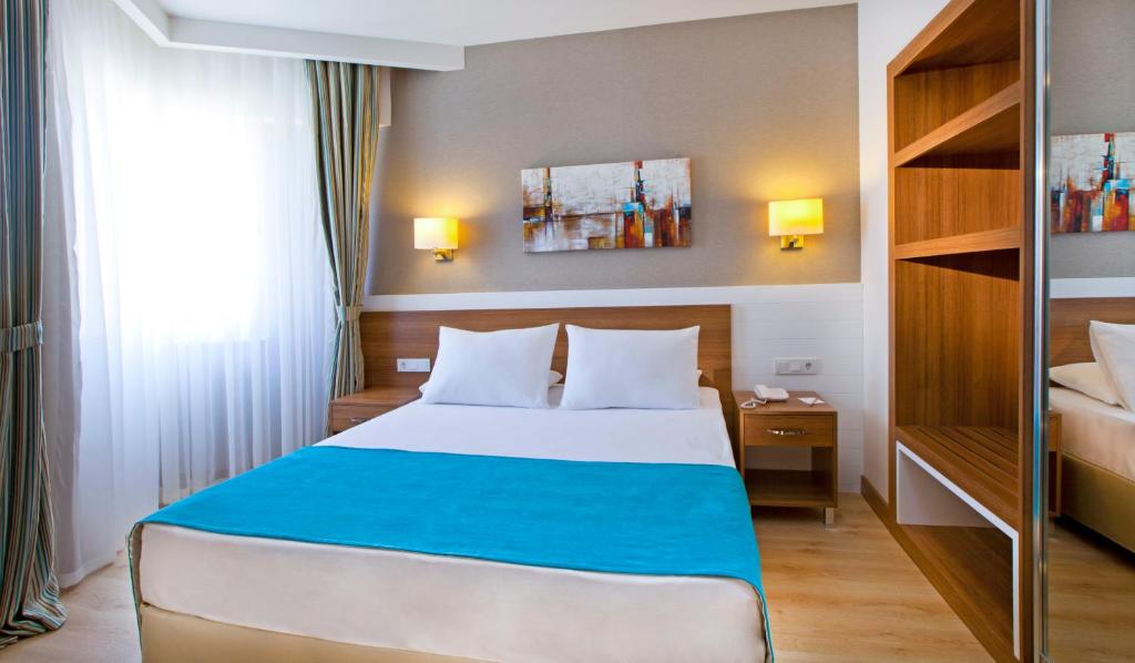 Mūsdienīga viesnīcas numura iekārtojums ar gultu, siltām krāsām un elegantu apgaismojumu, kas rada patīkamu atmosfēru. Numurā ir arī koka plaukti un mākslas darbs uz sienas.