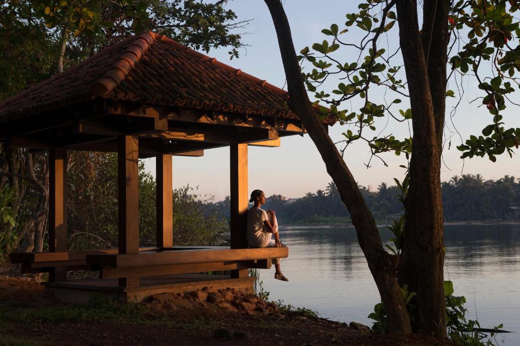Koka paviljons ar sarkanu jumtu pie ūdens, kur cilvēks sēž un izskata ainavu. Apvienojums ar dabu un mierīgu atmosfēru.