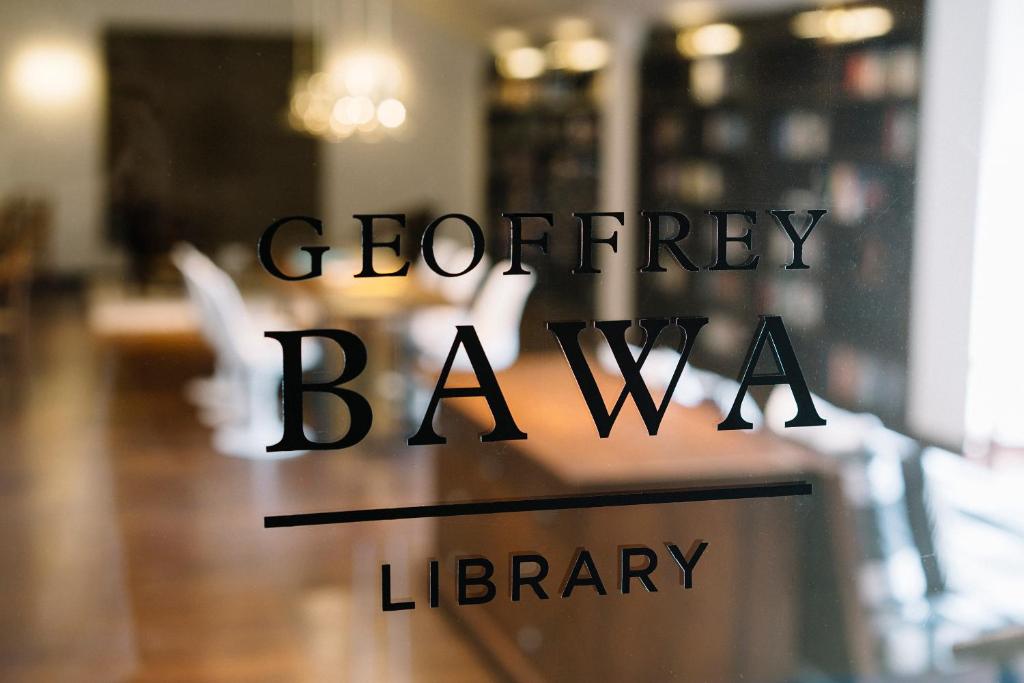 Geoffrey Bawa bibliotēkas nosaukums uz stikla durvīm, redzama moderna telpa ar koka grīdu un grāmatplauktiem fonā.