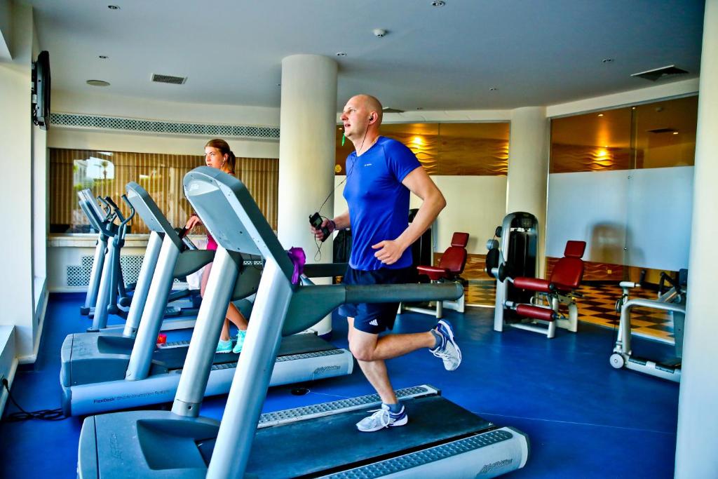 Vīrietis un sieviete trenējas uz skrejceliņiem modernā sporta zālē, kamēr televizors rāda programmu. Telpa ir gaiša un gaumīgi iekārtota.