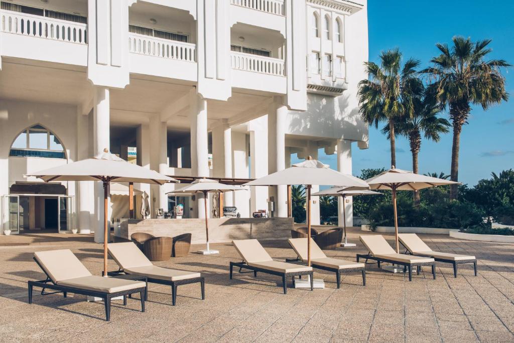 Saules gultas ar lietussargiem pie luksusa viesnīcas terases, ko ieskauj palmas un zila debesis. Ideāla vieta atpūtai un relaksācijai.