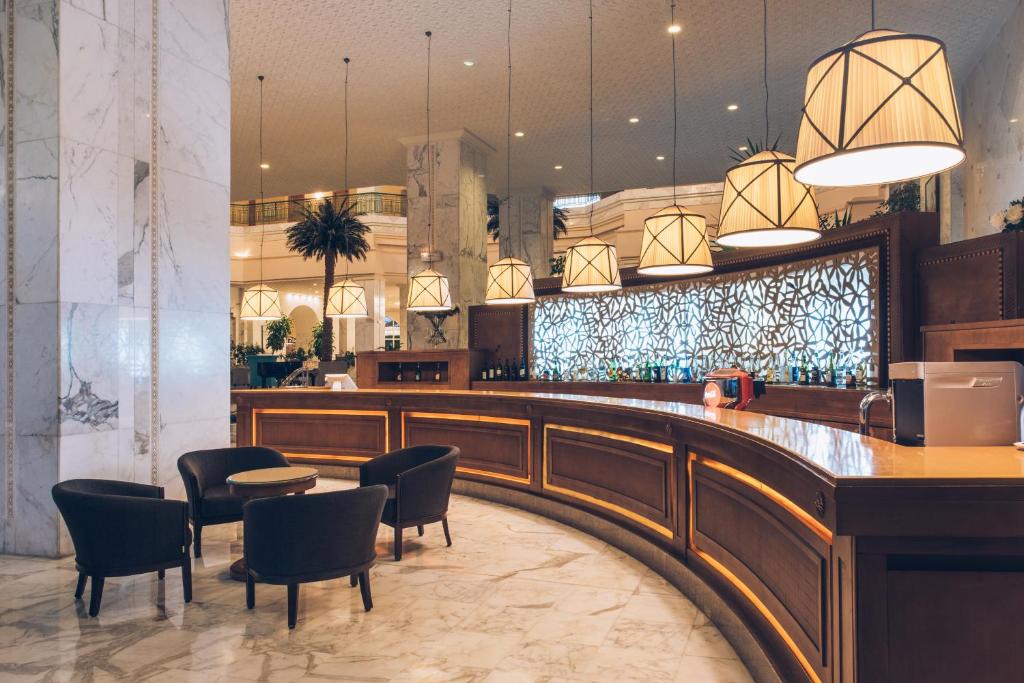 Elegants viesnīcas bārs ar marmorētām grīdām un stilīgām lampām, kas nodrošina siltu apgaismojumu. Bāra letes aizmugurē ir dzērienu plaukti, radot viesmīlīgu atmosfēru.