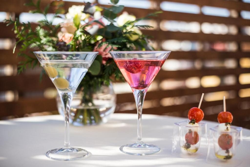 Divi kokteiļi glītās glāzēs, viens ar olīvām un otrs ar avenēm, kopā ar uzkodām, dekorēti ar ziediem.