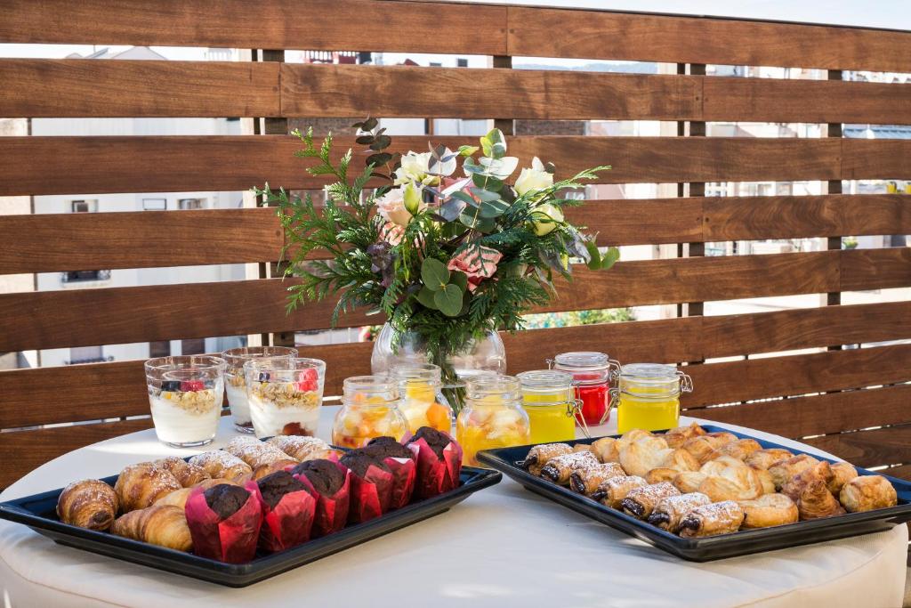 Svaiga brokastu izkārtojums uz balkona, kas ietver dažādus konditorejas izstrādājumus, augļu salātus un dzērienus, papildināts ar ziedu kompozīciju.
