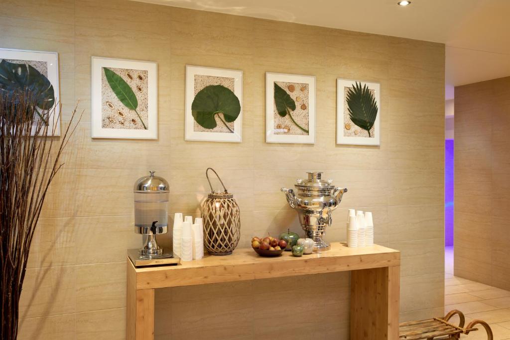 Moderns interjers ar dekoratīvām augu kompozīcijām uz sienas, zelta krāsas traukiem un dzērienu dozatoru uz koka galda.