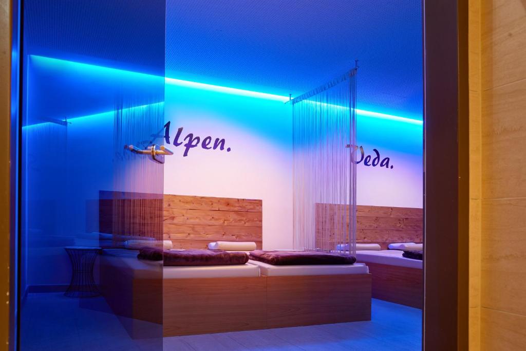 Mūsdienīga spa telpa ar relaksējošu zilu apgaismojumu un koka gultām, kas aicina uz atpūtu. Uz sienas redzami vārdi "Alpen" un "Deda".