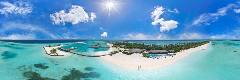 Tropu pludmale ar baltu smilšu un kristāldzidru ūdeni, kur redzama viesnīca un palmas, zem saulainas debess.