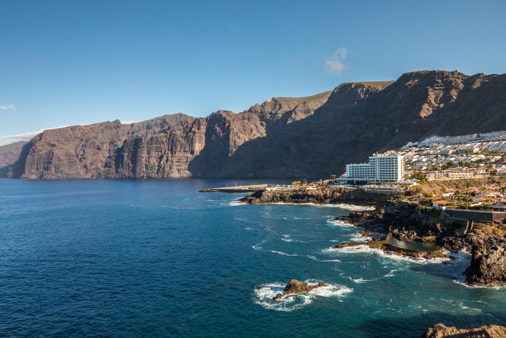 Krāšņa ainava ar klintīm, kas paceļas virs zilo ūdeņu, piekrastē redzama viesnīca un nelielas apdzīvotas vietas.