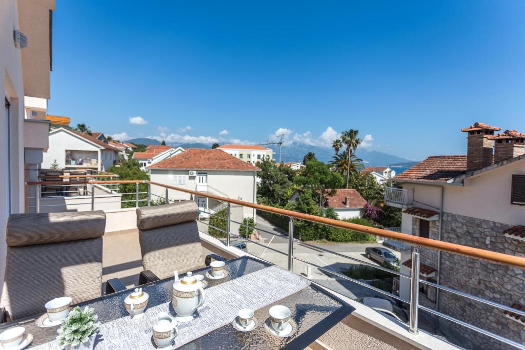 Saulains balkons ar skatu uz pilsētu, kur ir kafijas komplekts un ērtas krēslas. Fons rāda mājas un kalnus, radot mierīgu atmosfēru.
