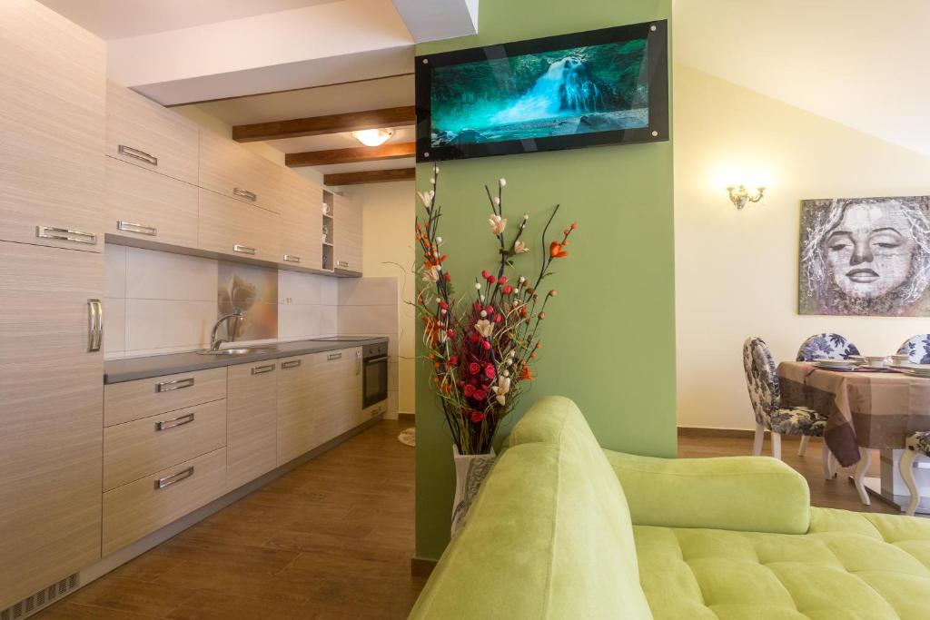 Mūsdienīga virtuve ar gaišām koka mēbelēm un zaļām sienām, kurā redzams televizors un ziedu kompozīcija. Ēdamzona ar stilīgām krēsliem un mākslas darbiem.