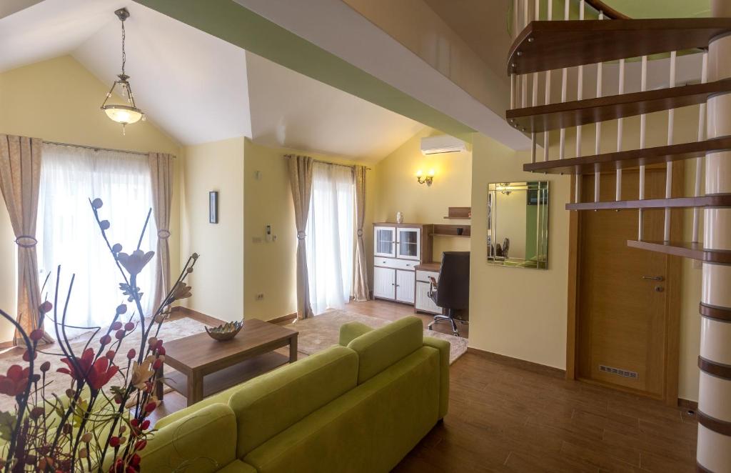 Mūsdienīgs viesistabas interjers ar zaļu dīvānu, koka galdu un kāpnēm, kas ved uz otro stāvu. Telpā ir lieli logi ar gaismas aizkariem un moderns mēbeļu komplekts.