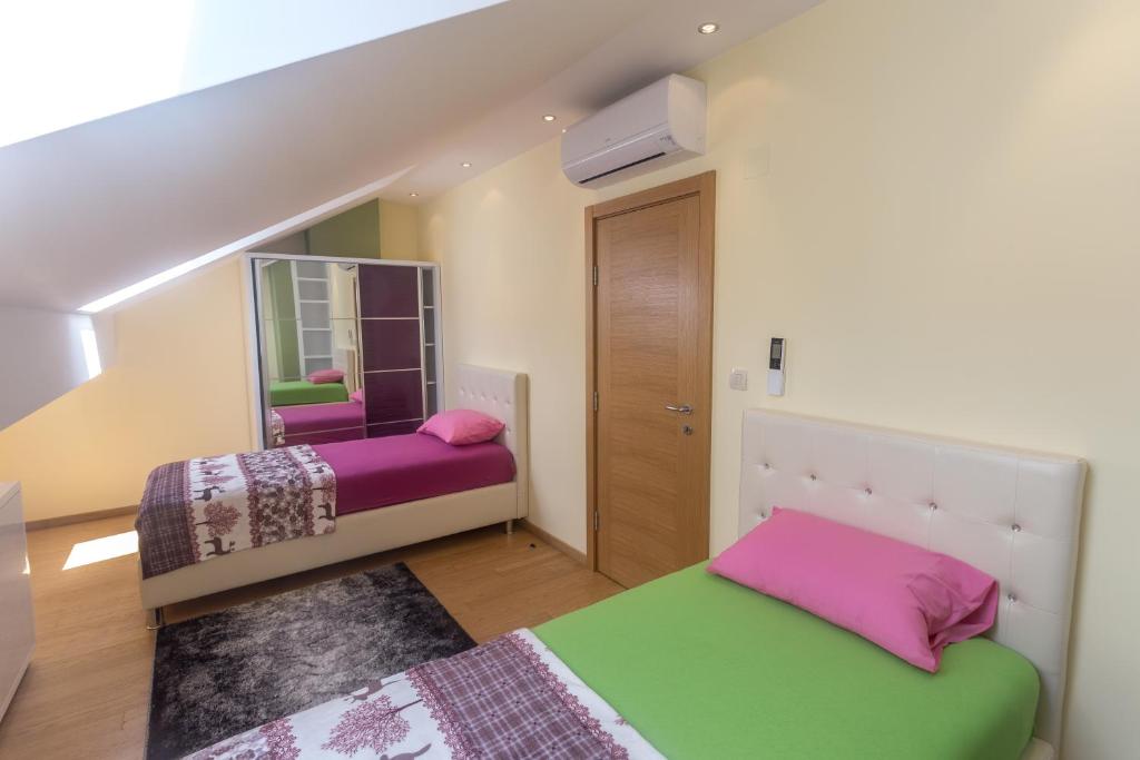 Mūsdienīga guļamistaba ar divām gultām, spilveniem rozā un zaļā krāsā, un lielu spoguli. Telpā ir gaišas sienas un koka grīda, radot patīkamu atmosfēru.