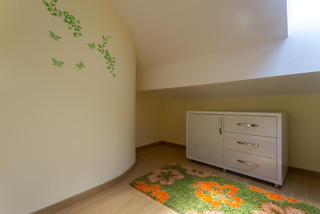 Gaiša telpa ar zaļiem tauriņu uz sienas, stilīgs balts kumode un krāsains paklājs ar ziedu motīviem.