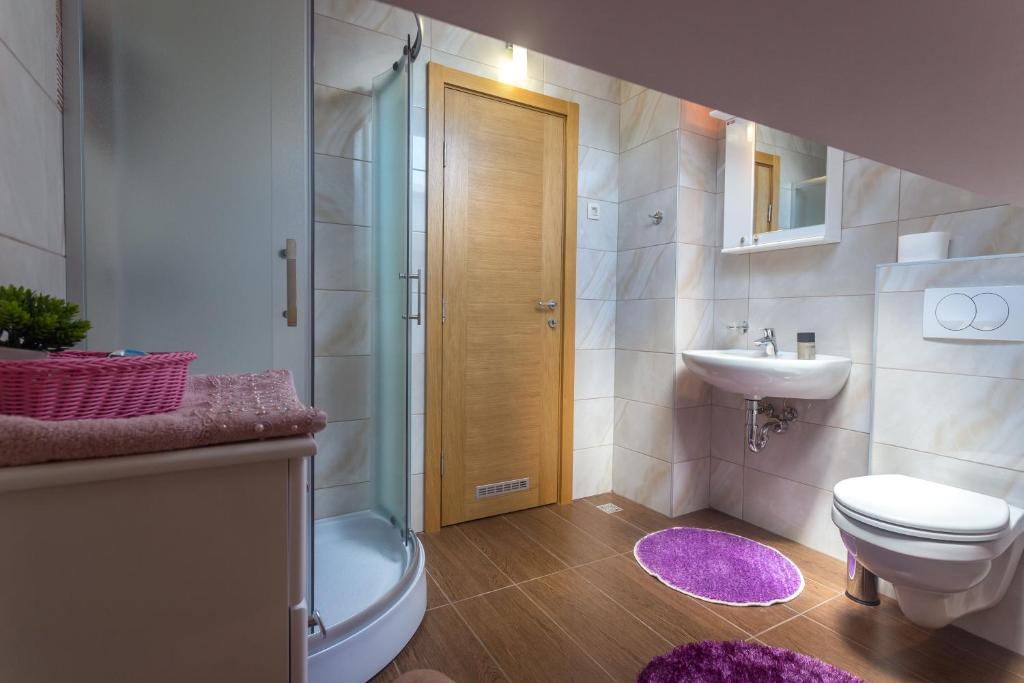 Mūsdienīgs vannas istabas interjers ar dušu, gaišām flīzēm un koka durvīm, papildināts ar rozā paklājiem un dekoratīvu augu.