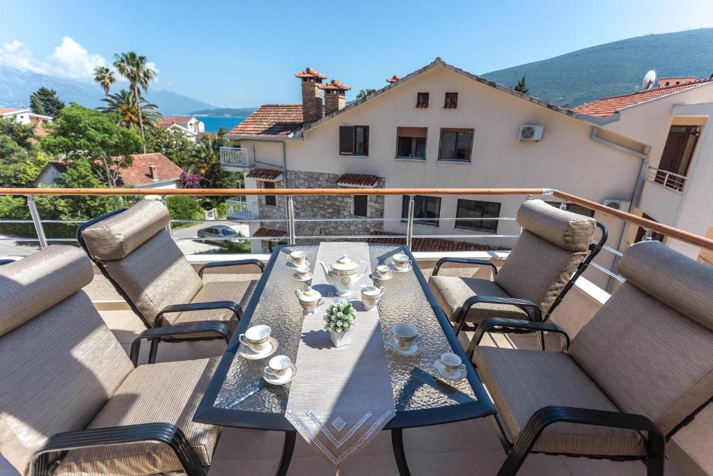 Terase ar skatu uz jūru, kur uz galda ir kafijas komplekts un četri ērti krēsli. Apkārt ir mājas ar sarkaniem jumtiem un zaļām palodzēm.