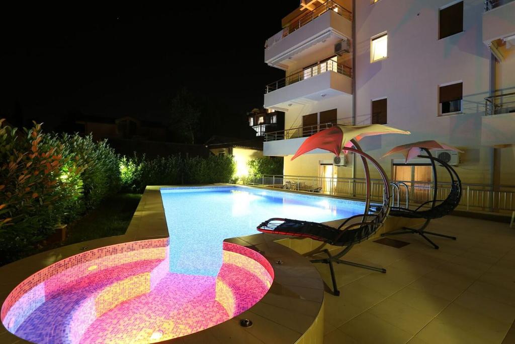 Mūsdienīga nakts ainava ar krāšņu baseinu un hidromasāžas vannu, apgaismojumu un atpūtas krēsliem, kas rada relaksējošu atmosfēru.