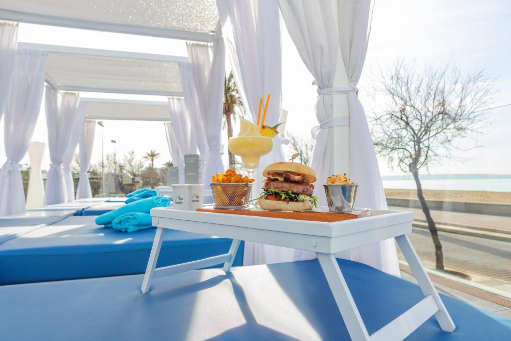 Piknika galds ar burgeru, frī kartupeļiem un kokteili pludmales kafejnīcā, ar baltām aizkariem un zilo dīvānu fonā. Ideāla atpūtas atmosfēra pie jūras.