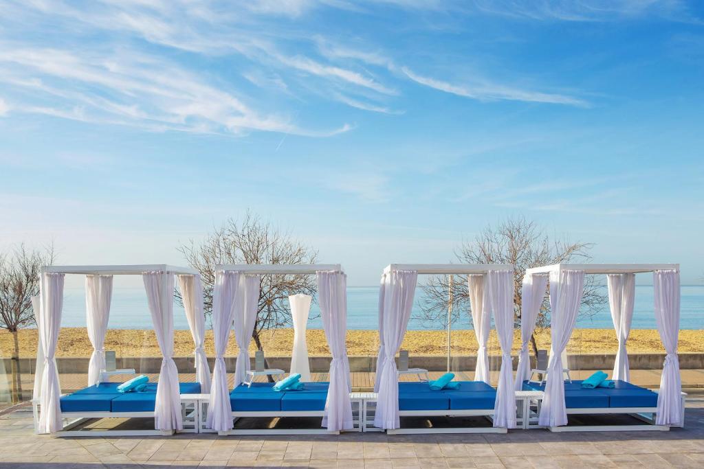 Pludmales gultas ar baltām aizkarām un zilu spilvenu rindu pie jūras, ar skatu uz smilšu pludmali un zilo debesu fonu.