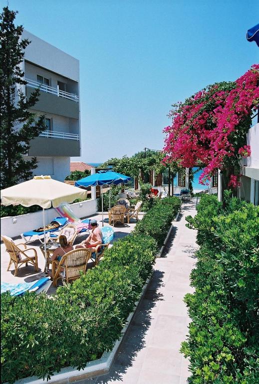 Atpūtas zona ar saules krēsliem, sauļošanās lietussargiem un ziedošiem augiem, kas atrodas pie jūras. Skats uz viesnīcu un zilo debesu fonu.