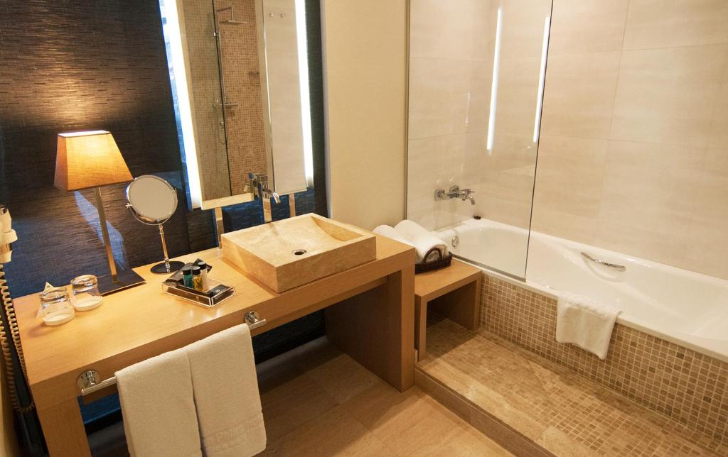 Mūsdienīgs vannas istabas interjers ar koka izlietni, gaismas spoguli un vannu. Telpa ir gaiša un gaumīga, nodrošinot relaksējošu atmosfēru.